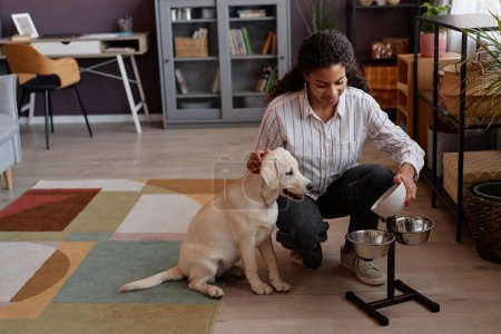 Photo for Full length portrait of smiling black woman feeding puppy at home and using stand with metal bowls - Royalty Free Image