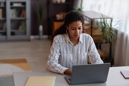 Téléchargez les photos : Vue de face portrait de jeune femme noire utilisant un ordinateur portable tout en travaillant à la maison dans un cadre confortable, espace de copie - en image libre de droit