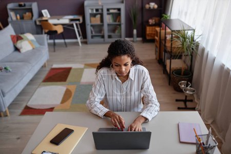 Foto de Vista de ángulo alto en la mujer joven negra usando el ordenador portátil mientras que trabaja en la oficina en el ajuste acogedor, espacio de copia - Imagen libre de derechos