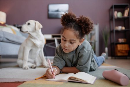 Foto de Retrato de niña preadolescente negro escribiendo en el diario acostado en el suelo en casa con perro en el fondo, espacio de copia - Imagen libre de derechos