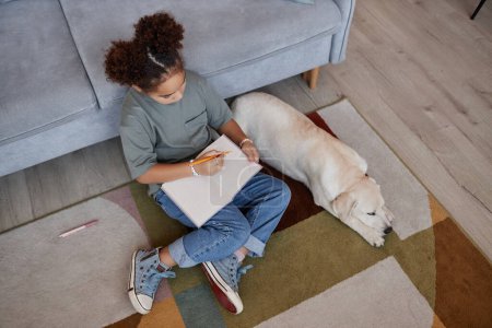 Téléchargez les photos : Vue de dessus à noir adolescent fille écriture dans le journal tandis que assis sur le sol à la maison avec chien blanc, espace de copie - en image libre de droit