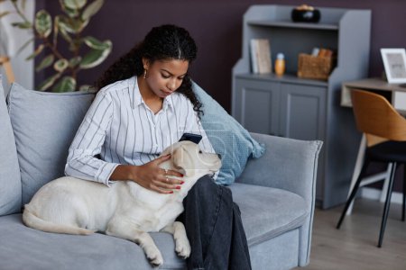 Téléchargez les photos : Portrait de jeune femme noire câlinant avec chiot mignon sur le canapé à la maison, chien mettant la tête dans les genoux avec amour - en image libre de droit