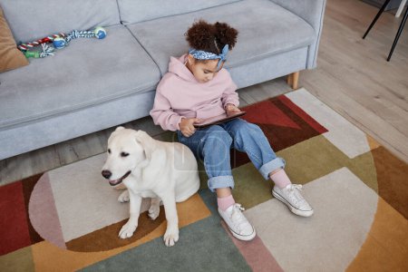 Téléchargez les photos : Portrait à angle élevé de mignonne fille noire en utilisant une tablette assise sur le sol à la maison avec un chien mignon, espace de copie - en image libre de droit