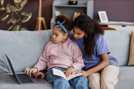 Foto de Retrato de vista lateral de una joven negra enseñando a su hija usando un portátil mientras están sentados juntos en un sofá - Imagen libre de derechos