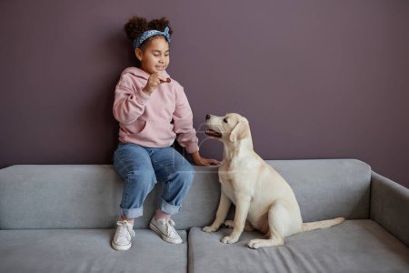 Téléchargez les photos : Portrait complet de petite fille noire jouant avec le chien à l'intérieur contre le mur violet, espace de copie - en image libre de droit