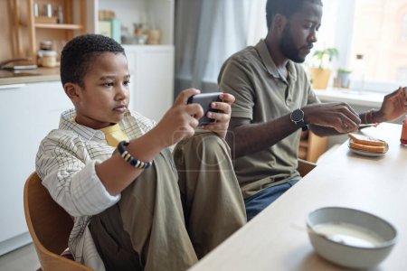 Téléchargez les photos : Portrait d'un adolescent afro-américain utilisant un smartphone à la table de cuisine pendant le petit déjeuner avec un espace de copie familiale - en image libre de droit