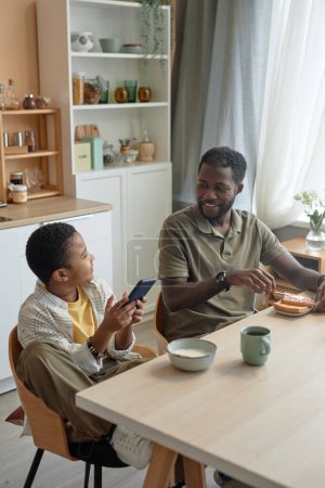 Téléchargez les photos : Portrait vertical du père et du fils afro-américains souriants assis à la table de la cuisine ensemble pendant le petit déjeuner le matin - en image libre de droit
