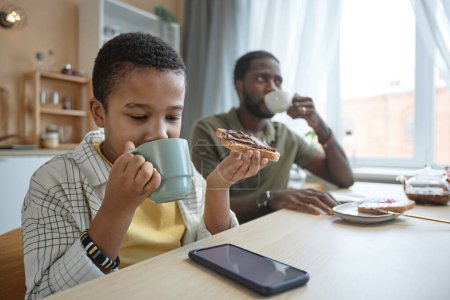 Téléchargez les photos : Portrait d'un garçon afro-américain prenant son petit déjeuner à la table de la cuisine en famille et regardant un écran de smartphone regarder des vidéos en ligne - en image libre de droit