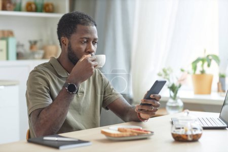 Téléchargez les photos : Portrait de l'homme afro-américain adulte utilisant un smartphone tout en prenant le petit déjeuner à la table de cuisine avec ordinateur portable - en image libre de droit