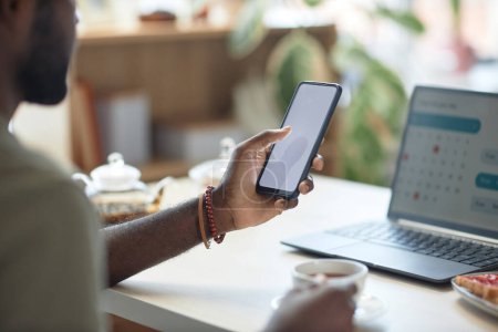 Téléchargez les photos : Gros plan de l'homme afro-américain méconnaissable utilisant un smartphone avec écran blanc vierge tout en travaillant à la table de cuisine - en image libre de droit