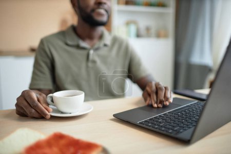 Téléchargez les photos : Gros plan de l'homme afro-américain utilisant un ordinateur portable et travaillant à la table de cuisine pendant l'espace de copie du petit déjeuner - en image libre de droit
