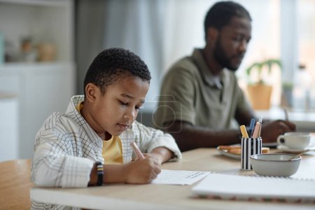 Téléchargez les photos : Portrait latéral d'un garçon afro-américain faisant ses devoirs à la table de cuisine avec son père travaillant dans un espace de copie de fond - en image libre de droit
