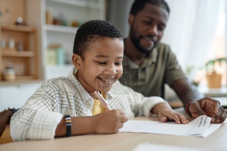 Téléchargez les photos : Portrait d'un garçon noir souriant faisant ses devoirs à la table de cuisine avec son père aidant à copier l'espace - en image libre de droit