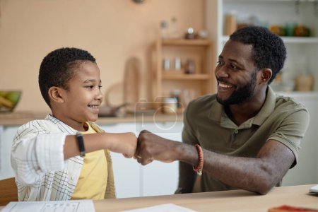 Téléchargez les photos : Portrait du père et du fils afro-américains heureux coup de poing tout en faisant des devoirs ensemble et en célébrant le succès dans l'étude - en image libre de droit