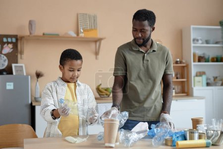 Téléchargez les photos : Portrait de la famille afro-américaine de père et fils triant déchets et plastique ensemble dans la cuisine de la maison - en image libre de droit
