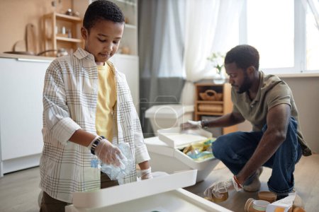 Portrait of young Black boy sorting waste at home with father and putting plastic bottle into recycling bin copy space