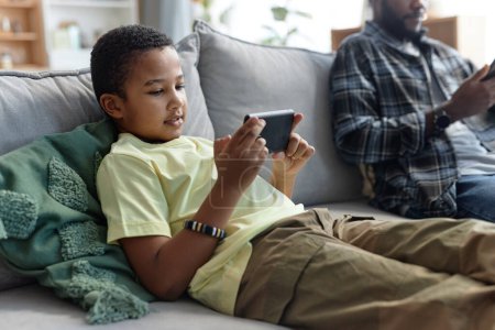 Foto de Retrato de un adolescente afroamericano usando un teléfono inteligente y viendo videos en línea mientras se relaja en el sofá en casa - Imagen libre de derechos