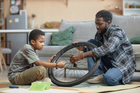 Photo for Side view portrait of African American father and son repairing bicycle wheel together sitting on floor at home - Royalty Free Image