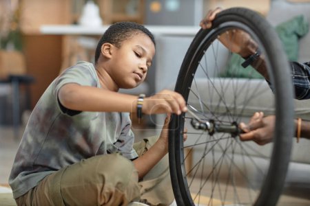 Side view portrait of young Black boy repairing bicycle wheel with father sitting on floor at home