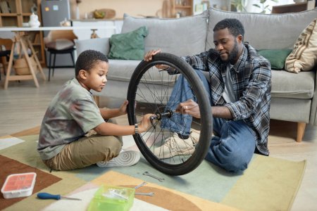Photo for Side view portrait of Black young father and son fixing bicycle wheels together at home - Royalty Free Image