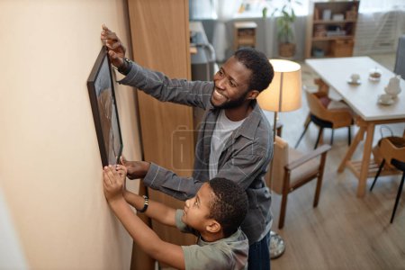 Foto de Vista lateral retrato de sonriente Negro padre e hijo colgando cuadro en la pared juntos y sonriendo felizmente - Imagen libre de derechos