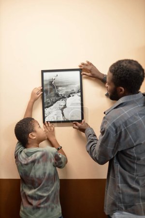 Téléchargez les photos : Vue arrière verticale du père et du fils accrochant l'image sur le mur ensemble dans la nouvelle maison - en image libre de droit