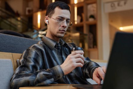 Foto de Retrato de un joven enfocado que usa gafas y bebe café mientras trabaja con una computadora portátil en el salón de oficina o en el vestíbulo del hotel - Imagen libre de derechos
