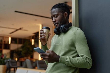 Foto de Retrato de vista lateral mínimo del joven negro usando un teléfono inteligente apoyado en la pared en el salón de oficina y bebiendo espacio para copias de café - Imagen libre de derechos