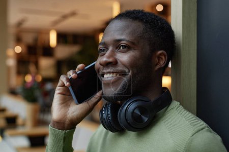 Photo for Side view portrait of Black young man speaking on phone and smiling in modern office lounge copy space - Royalty Free Image