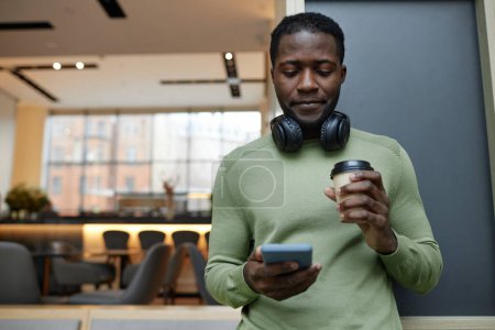 Foto de Retrato de cintura mínima hacia arriba del joven afroamericano usando un teléfono inteligente apoyado en la pared en el salón de oficina y bebiendo espacio para copiar café - Imagen libre de derechos