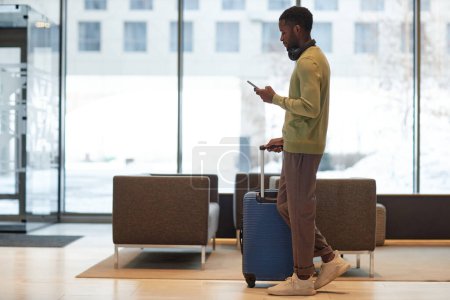 Foto de Retrato de cuerpo entero con vista lateral del hombre afroamericano con equipaje caminando por el vestíbulo del hotel y usando la aplicación de taxi para llamadas de teléfonos inteligentes - Imagen libre de derechos