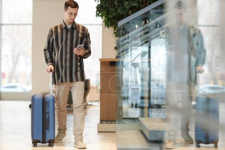 Téléchargez les photos : Portrait complet d'un jeune homme moderne avec valise marchant vers la caméra dans le hall de l'hôtel et utilisant un smartphone avec espace de copie d'application de taxi - en image libre de droit