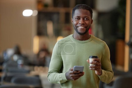 Téléchargez les photos : Portrait d'un homme afro-américain souriant tenant une tasse en papier avec café et smartphone dans un espace de copie de café de bureau - en image libre de droit