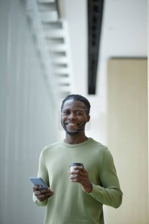 Foto de Retrato vertical mínimo del hombre afroamericano sonriente sosteniendo la taza de café y el teléfono de pie en el interior del edificio de oficinas limpio - Imagen libre de derechos