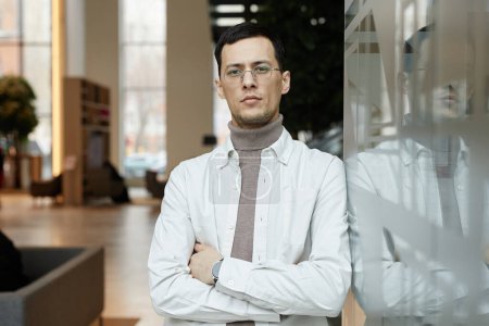 Photo for Minimal waist up portrait of confident Asian man wearing glasses leaning on glass wall in office building and looking at camera - Royalty Free Image