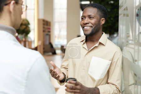 Photo for Waist up portrait of smiling Black young man chatting with colleague during break in office building and gesturing copy space - Royalty Free Image