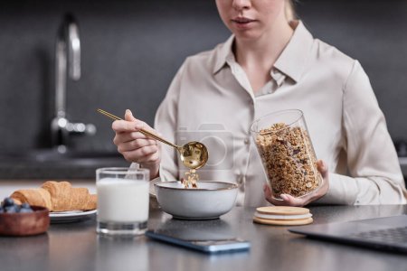 Téléchargez les photos : Gros plan de jeune femme profitant d'un petit déjeuner sain le matin versant granola dans un bol avec cuillère dorée, espace de copie - en image libre de droit
