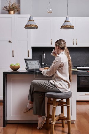 Foto de Retrato vertical de longitud completa de mujer joven usando el ordenador portátil en la mesa de la cocina en el interior del hogar - Imagen libre de derechos