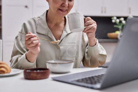 Téléchargez les photos : Gros plan de la femme âgée moderne utilisant un ordinateur portable tout en appréciant le petit déjeuner à la maison - en image libre de droit