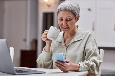 Téléchargez les photos : Portrait de femme âgée moderne utilisant un smartphone au petit déjeuner le matin et buvant du café, espace de copie - en image libre de droit