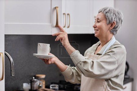 Foto de Retrato de vista lateral de mujer mayor sonriente abriendo gabinetes de cocina en casa y sosteniendo la taza de té, espacio para copiar - Imagen libre de derechos