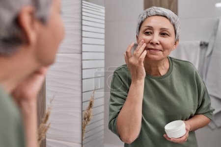 Téléchargez les photos : Portrait d'une femme asiatique âgée souriante regardant dans un miroir et utilisant de la crème visage - en image libre de droit