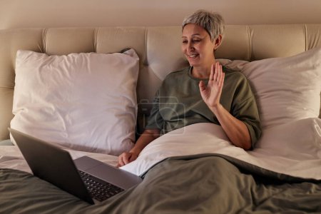 Retrato de una mujer mayor sonriente usando video chat y saludando a la cámara mientras está acostada en la cama en casa, espacio para copiar