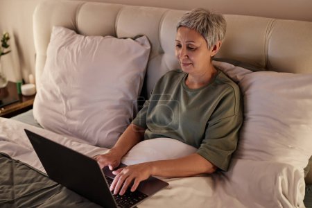 Retrato de ángulo alto de la mujer mayor asiática usando el ordenador portátil mientras que miente en la cama cómoda en el país
