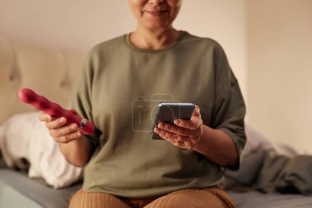 Photo for Close up of senior woman holding sex toy and smartphone while sitting on bed at home - Royalty Free Image