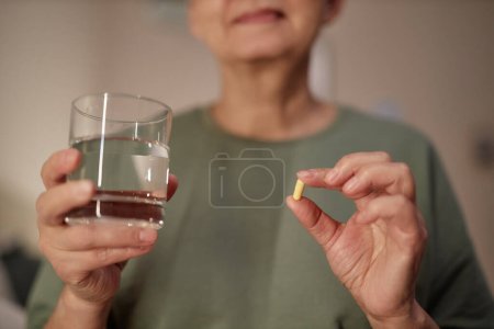 Photo for Close up of senior woman holding glass of water and taking medication at night, copy space - Royalty Free Image