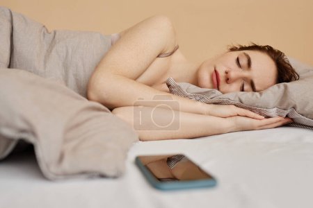 Foto de Retrato mínimo de una mujer joven acostada en una cama cómoda y durmiendo con un smartphone cerca, espacio para copiar - Imagen libre de derechos