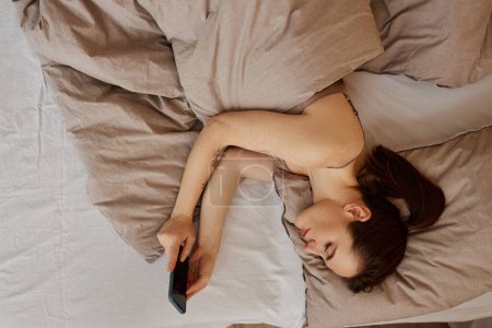 Mínimo retrato de vista superior de una mujer joven acostada en una cama cómoda y usando las redes sociales de desplazamiento de teléfonos inteligentes, espacio para copiar