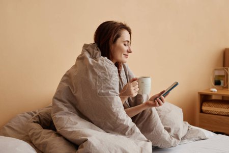 Mínimo retrato de vista lateral de mujer joven cubierta de manta acogedora disfrutando de un fin de semana relajante en casa y sosteniendo el teléfono, espacio para copiar