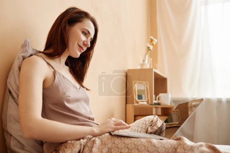 Retrato de vista lateral de una mujer joven usando un portátil en la cama en casa y sonriendo, espacio para copiar